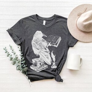 May include: A dark gray t-shirt with a white graphic of a raven perched on a book with a quill and inkwell. The raven is holding a book open with text visible.