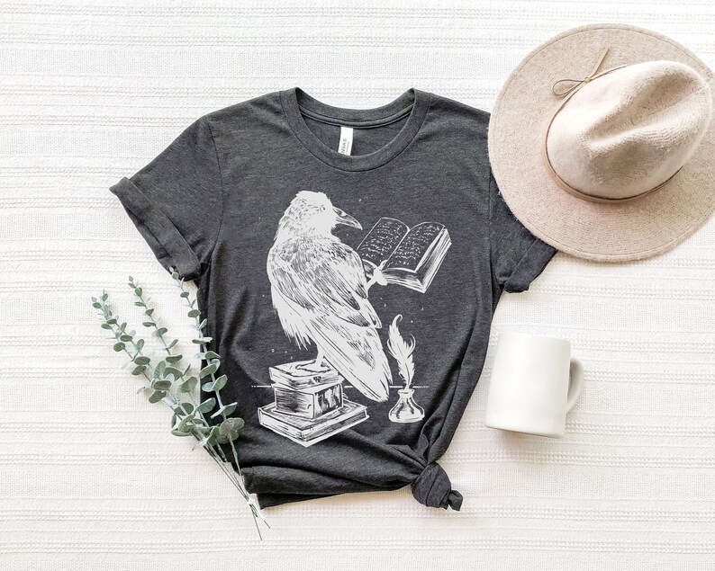 May include: A dark gray t-shirt with a white graphic of a raven perched on a book with a quill and inkwell. The raven is holding a book open with text visible.
