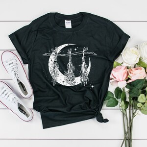 Pu&ograve; includere: T-shirt nero con un disegno a mezzaluna bianca e mazzi di erbe appesi. L'immagine include scarpe da ginnastica bianche e un bouquet di rose rosa e bianche. La maglietta &egrave; annodata in vita.