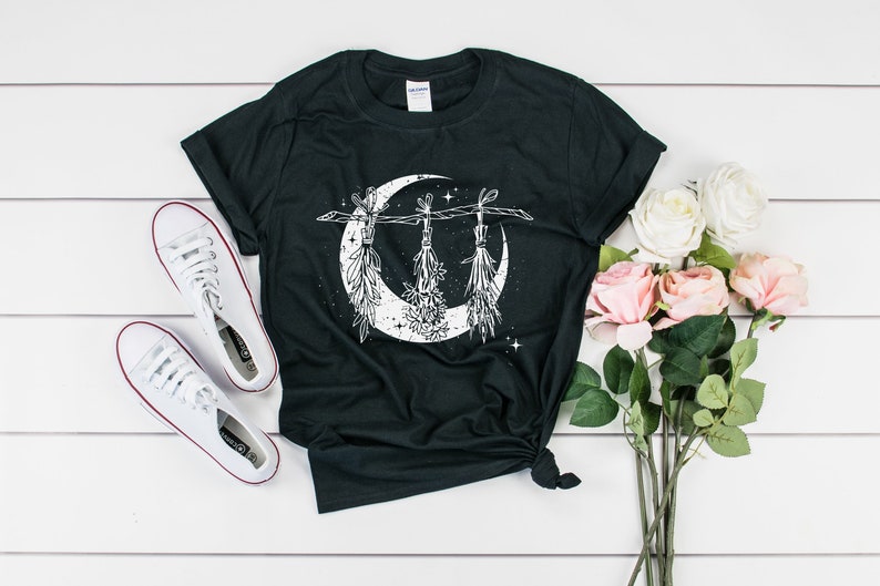 Pu&ograve; includere: T-shirt nero con un disegno a mezzaluna bianca e mazzi di erbe appesi. L'immagine include scarpe da ginnastica bianche e un bouquet di rose rosa e bianche. La maglietta &egrave; annodata in vita.