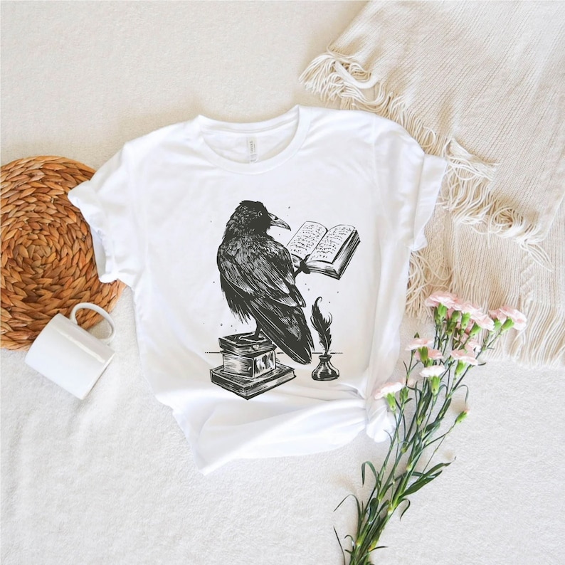 May include: White t-shirt with a black and white illustration of a raven perched on a stack of books, holding an open book in its talons. A quill pen and inkwell are beside the raven.