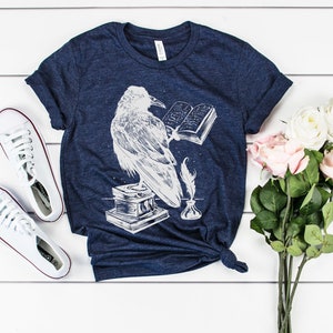 May include: A navy blue t-shirt with a white graphic of a raven reading a book, a quill pen, and an inkwell. The t-shirt is laid flat on a white wooden surface with a bouquet of pink and white roses and a pair of white sneakers.