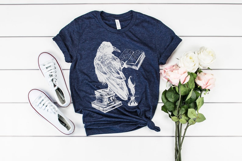 May include: A navy blue t-shirt with a white graphic of a raven reading a book, a quill pen, and an inkwell. The t-shirt is laid flat on a white wooden surface with a bouquet of pink and white roses and a pair of white sneakers.