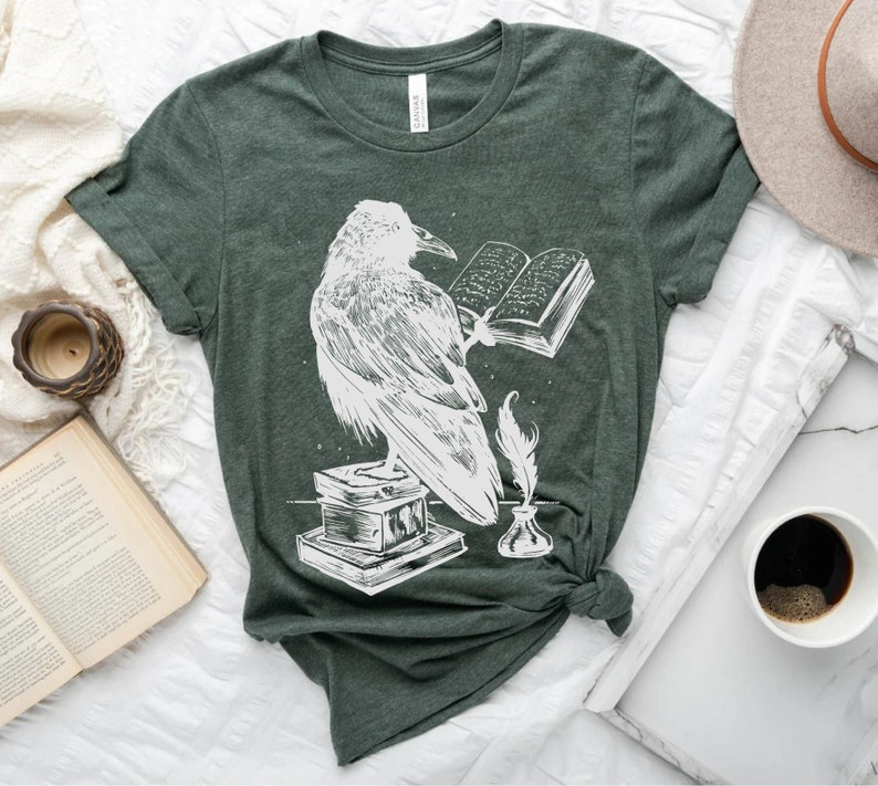 May include: A dark green t-shirt with a white graphic of a raven perched on a stack of books, holding an open book in its talons. A quill pen and inkwell are beside the raven.