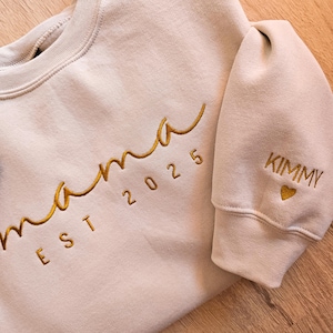 Könnte beinhalten: Ein beiger Pullover mit goldener Stickerei, auf dem vorne "mama est 2025" und auf dem Ärmel "KIMMY" mit einem Herz steht.