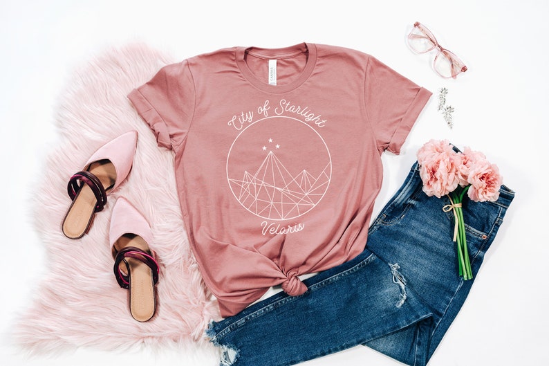 May include: A dusty rose t-shirt with a white graphic design that says "City of Starlight Velaris". The graphic features a geometric mountain range with stars inside a circle.