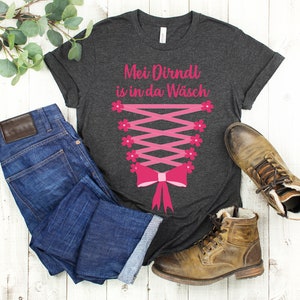 May include: Dark gray t-shirt with a pink corset design and the text "Mei Dirndl is in da W&auml;sch". The shirt is paired with blue jeans and brown leather boots, creating a casual, everyday look.