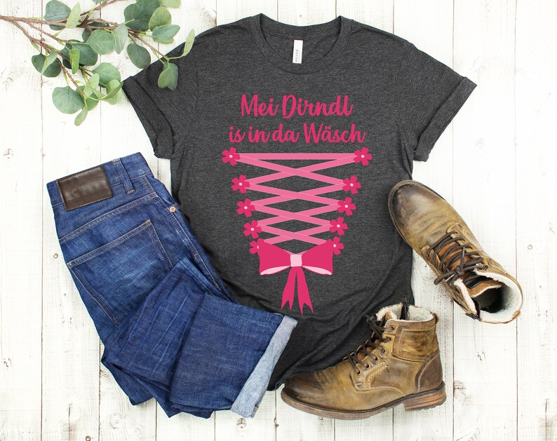 May include: Dark gray t-shirt with a pink corset design and the text "Mei Dirndl is in da W&auml;sch". The shirt is paired with blue jeans and brown leather boots, creating a casual, everyday look.