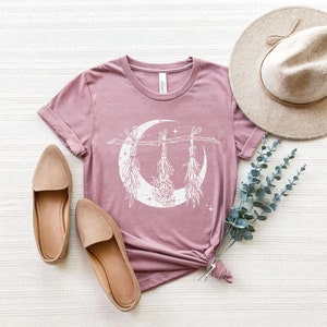 Pu&ograve; includere: T-shirt malva con grafica bianca a forma di luna crescente e mazzi di fiori appesi. L'immagine include ballerine beige, un cappello a tesa larga e rametti di verde. La maglietta &egrave; annodata in vita.