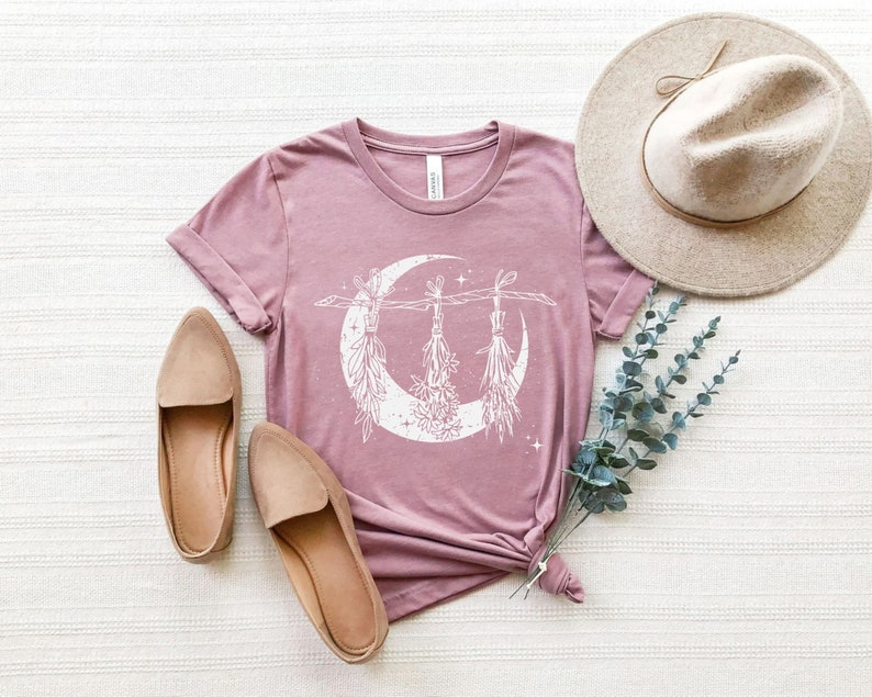 Pu&ograve; includere: T-shirt malva con grafica bianca a forma di luna crescente e mazzi di fiori appesi. L'immagine include ballerine beige, un cappello a tesa larga e rametti di verde. La maglietta &egrave; annodata in vita.