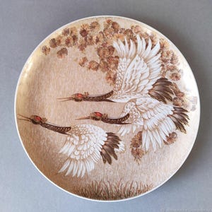 May include: A round decorative plate featuring a detailed illustration of three cranes in flight. The cranes have white feathers with brown accents and red crests. The background is a beige color with floral and grass details.