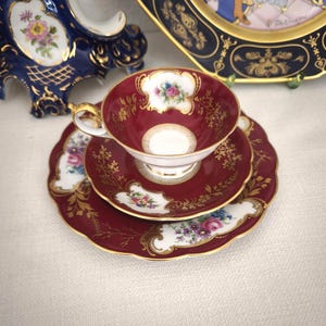 Może przedstawiać: Bordowy porcelanowy zestaw do herbaty, składający się z filiżanki, spodka i talerza. Zestaw posiada złote filigranowe i kwiatowe akcenty. Filiżanka i spodek są umieszczone na talerzu. Klasyczny design.