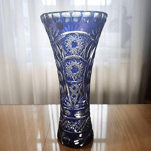 Peut inclure: Un vase en verre bleu cobalt, orné de motifs complexes en forme d'étoiles et de géométries taillées. Le vase a un sommet évasé et une base ronde. Le design en verre taillé reflète la lumière, créant un effet scintillant. Le vase est probablement décoratif.