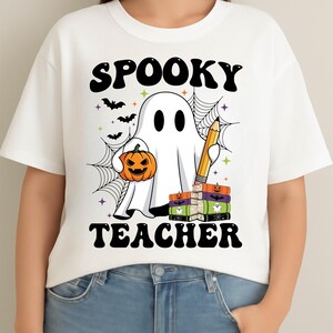 May include: White t-shirt with a Halloween-themed graphic. The design features a ghost holding a jack-o'-lantern and a pencil, with the words "SPOOKY TEACHER" in black. Bats and a spiderweb complete the design.