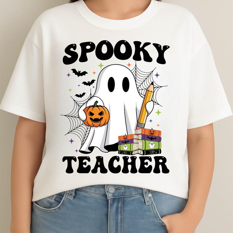 May include: White t-shirt with a Halloween-themed graphic. The design features a ghost holding a jack-o'-lantern and a pencil, with the words "SPOOKY TEACHER" in black. Bats and a spiderweb complete the design.