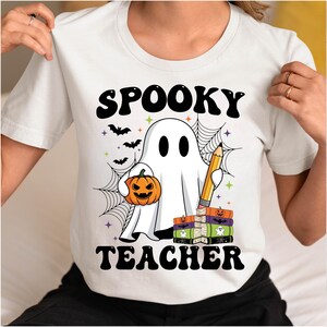 May include: White t-shirt with a Halloween-themed graphic. The design features a ghost holding a pumpkin and a pencil, with the words "Spooky Teacher" in black. Bats and a spiderweb complete the design.