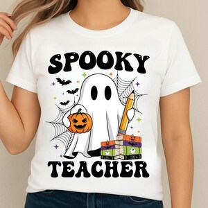 May include: White t-shirt with a Halloween-themed graphic. The design features a cartoon ghost holding a pumpkin and a pencil, with the words "Spooky Teacher" in black. Bats, spiderwebs, and stars complete the design.
