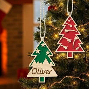 K&ouml;nnte beinhalten: Zwei h&ouml;lzerne Weihnachtsbaumschmuckst&uuml;cke, die an einem Weihnachtsbaum h&auml;ngen. Das Ornament links ist gr&uuml;n mit wei&szlig;en Sternen und dem Namen "Oliver" darauf geschrieben. Das Ornament rechts ist rot mit wei&szlig;en Sternen und wei&szlig;en Linien.