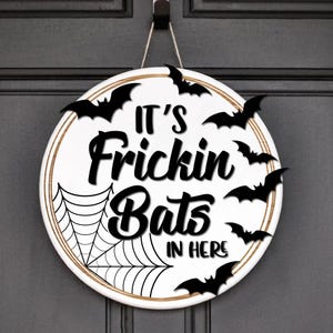 May include: White round door hanger with a black spiderweb and bats. The text "It's Frickin Bats In Here" is written in black.