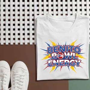 May include: White t-shirt with the words "BENITO BOWL ENERGY" in blue and red, with a football and Puerto Rican flag design. The text is surrounded by yellow and blue lightning bolt graphics. A pair of white sneakers are in the foreground.