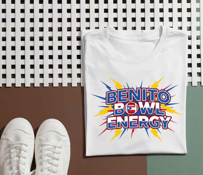 May include: White t-shirt with the words "BENITO BOWL ENERGY" in blue and red, with a football and Puerto Rican flag design. The text is surrounded by yellow and blue lightning bolt graphics. A pair of white sneakers are in the foreground.