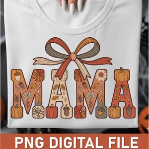 Pode incluir: Camiseta branca com a palavra "MAMA" em um design com tema de outono. As letras são decoradas com abóboras, bolotas e padrões florais em tons de laranja, marrom e creme. Um laço decorativo está posicionado acima da palavra.