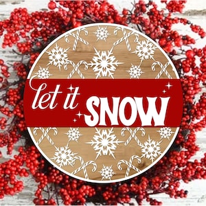 May include: A round wooden sign with the words "let it SNOW" in white on a red banner. The sign features snowflakes and candy canes on a light brown background, surrounded by a wreath of red berries.