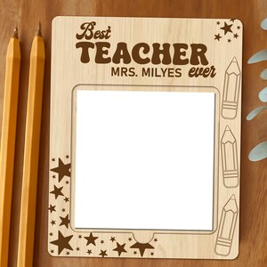 Op de afbeelding: Een houten frame met een witte inzet voor een foto. Het frame heeft de tekst "Best Teacher MRS. MILYES ever" en is versierd met sterren en potloden.