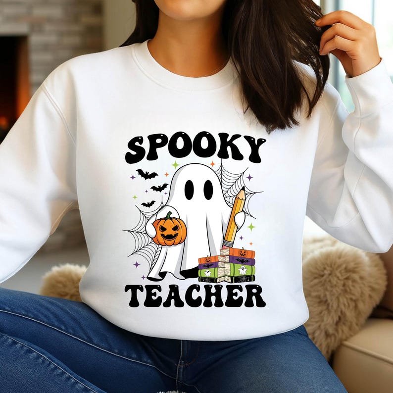 May include: White sweatshirt with a Halloween-themed graphic. The design features a ghost holding a pumpkin and a pencil, with the words "SPOOKY TEACHER" in black. Bats, spiderwebs, and books complete the design.