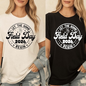 Könnte beinhalten: Zwei T-Shirts, eines weiß und eines schwarz, jeweils mit einem kreisförmigen Grafikdesign. Die Grafik lautet "LET THE GAME Field Day 2026 BEGIN" in einer stilisierten Schriftart, mit einem Friedenszeichen und Sternen. Die Shirts sind kurzärmelig.
