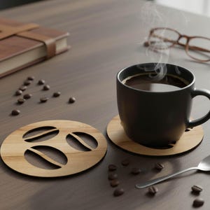 Puede incluir: Una taza de caf&eacute; negro con vapor ascendente, sobre un posavasos de madera con dise&ntilde;o de grano de caf&eacute;. Otro posavasos y granos de caf&eacute; dispersos. Tambi&eacute;n se ven una cuchara y gafas.