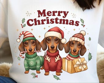 May include: White sweatshirt featuring a festive illustration of three dachshunds wearing Christmas sweaters and hats. The dogs are brown and black, with one holding a candy cane. Text reads "Merry Christmas" and "MERRY CHRISTMAS PNG PNG DIGITAL FILES".