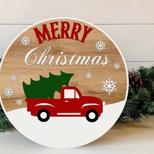 May include: Round wooden Christmas sign with a red truck carrying a green Christmas tree. The sign has the words "MERRY Christmas" in red and white, with white snowflakes. The background is a wood grain pattern.