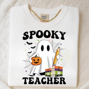 May include: White sweatshirt with a gold chain and the words "SPOOKY TEACHER" in black. A cartoon ghost holds a jack-o'-lantern and a pencil, with bats and spiderwebs. The sweatshirt has the "COMFORT COLORS" tag.