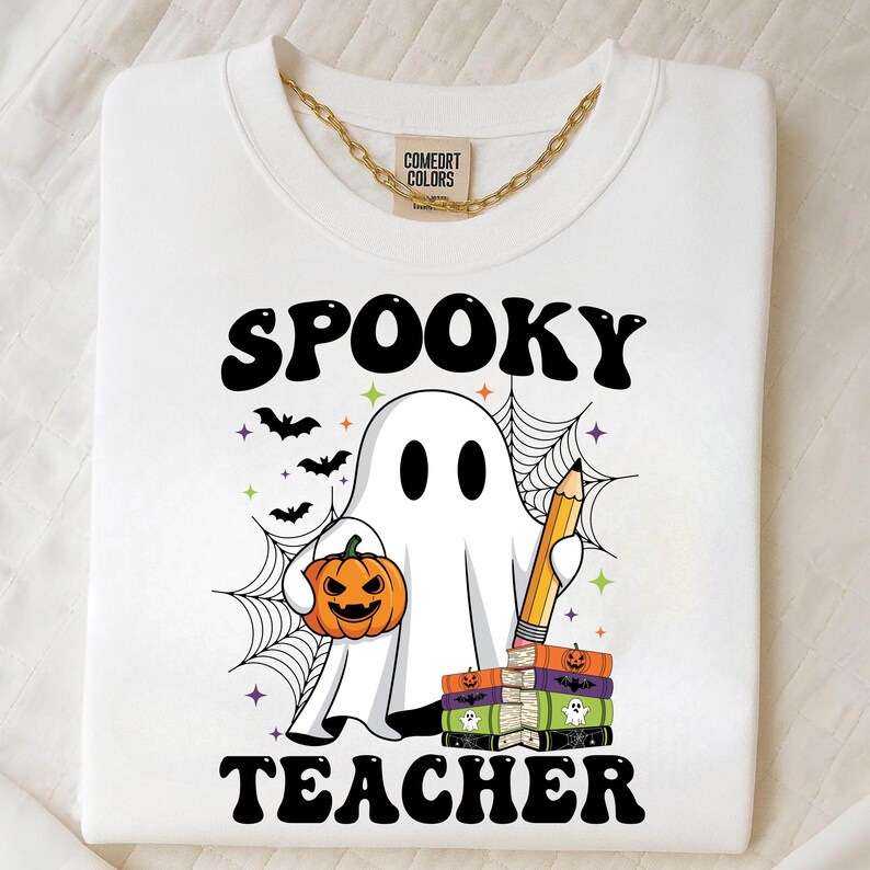 May include: White sweatshirt with a gold chain and the words "SPOOKY TEACHER" in black. A cartoon ghost holds a jack-o'-lantern and a pencil, with bats and spiderwebs. The sweatshirt has the "COMFORT COLORS" tag.