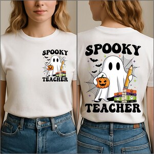 May include: White t-shirt with a Halloween-themed graphic. The design features a ghost holding a pumpkin and a pencil, with the words "Spooky Teacher" above and below the image. Bats and spiderwebs complete the design.