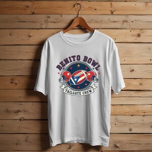 May include: White t-shirt with a graphic featuring a football, two helmets, and the text "Benito Bowl Tailgate Crew." The design incorporates red, white, and blue colors, with stars and a banner.