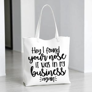 May include: White tote bag with black text that reads "Hey I found your nose it was in my business again".