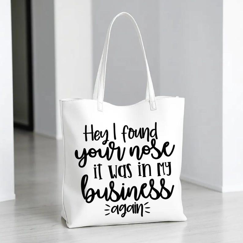 May include: White tote bag with black text that reads "Hey I found your nose it was in my business again".