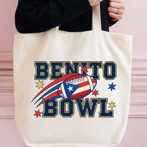 May include: Cream-colored tote bag with the words "BENITO BOWL" in navy blue block letters with gold trim. A football with the Puerto Rican flag design is in the center. Red, blue, and gold star graphics surround the text and football.