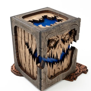 Op de afbeelding: Een houten kist met een gebeeldhouwd monstergezichtontwerp. De kist is bruin met een blauw interieur zichtbaar door de mond en bovenkant van het monster. De kist staat op een decoratieve houten basis.