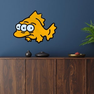 May include: A yellow cartoon fish with three eyes and a black outline is mounted on a dark blue wall. The fish has a grumpy expression. The wall art is above a dark wood cabinet with decorative items.