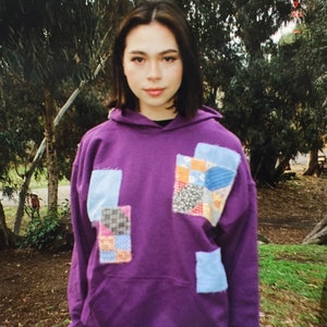 Pode incluir: Um moletom roxo com um design de patchwork na frente. Os patches são feitos de diferentes tecidos e cores, incluindo azul, vermelho e amarelo.