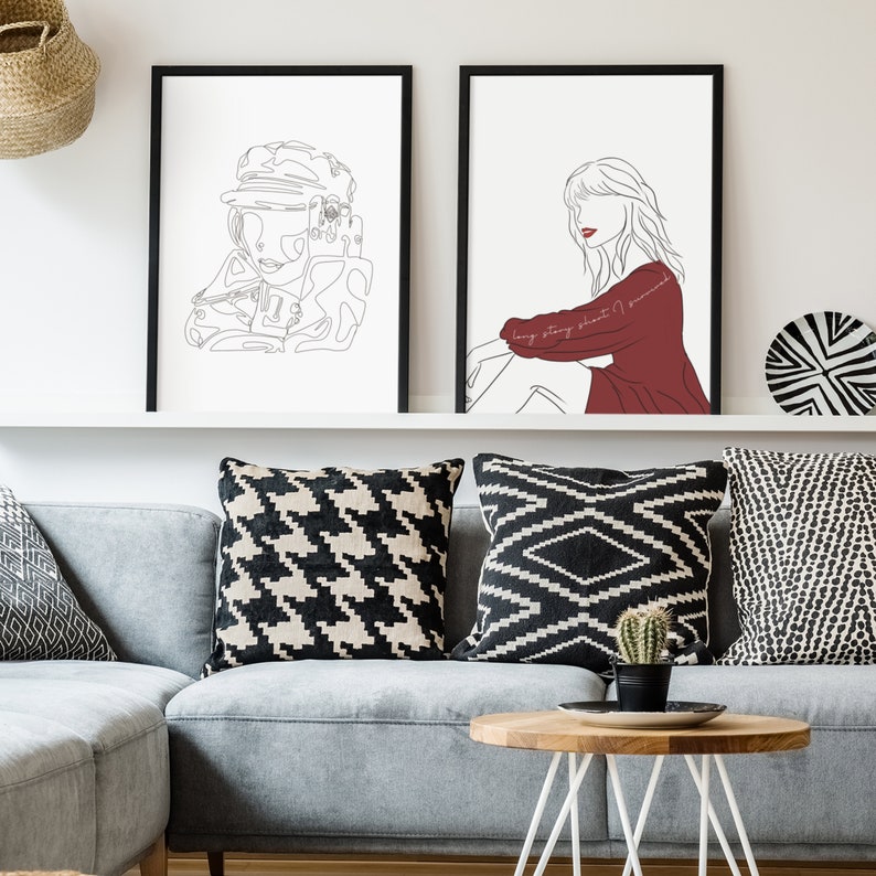 May include: Two framed art prints with black frames. One features a line drawing of a person in a hat, the other a person in a red top. The prints are displayed above a gray sofa with patterned pillows and a wooden coffee table.