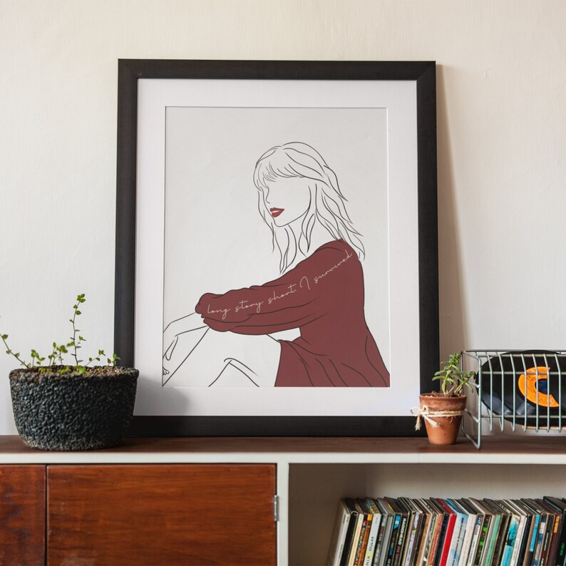 May include: Framed art print featuring a line drawing of a person in a maroon dress, with the text "long story short I survived" in cursive. The artwork is in a black frame and displayed on a wooden shelf.