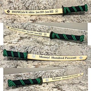 Könnte beinhalten: Ein Holzschwert mit einem schwarz-grünen Griff. Das Schwert ist mit dem Text "HM1(FMF) Julie M. Adkins June 2019 - June 2022" und "Money! Hundred Percent!" graviert.