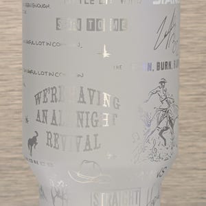 Może przedstawiać: Biały kubek ze srebrnym tekstem i grafiką. Kubek zawiera u g&oacute;ry imię "ZACH BRYAN", a także teksty piosenek i ilustracje kowboja na koniu, kapelusza i serca. Widoczne są r&oacute;wnież słowa "STRAIGHT OUTTA OKLAHOMA".