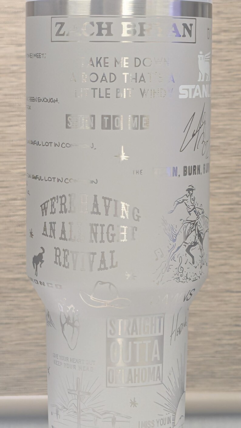 Może przedstawiać: Biały kubek ze srebrnym tekstem i grafiką. Kubek zawiera u g&oacute;ry imię "ZACH BRYAN", a także teksty piosenek i ilustracje kowboja na koniu, kapelusza i serca. Widoczne są r&oacute;wnież słowa "STRAIGHT OUTTA OKLAHOMA".