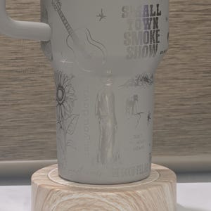 Może przedstawiać: Biały kubek z uchwytem, z wytrawionymi ilustracjami gitary, słonecznika, kowboja i konia. Widoczny jest napis "SMALL TOWN SMOKE SHOW". Kubek stoi na drewnianej podstawce.