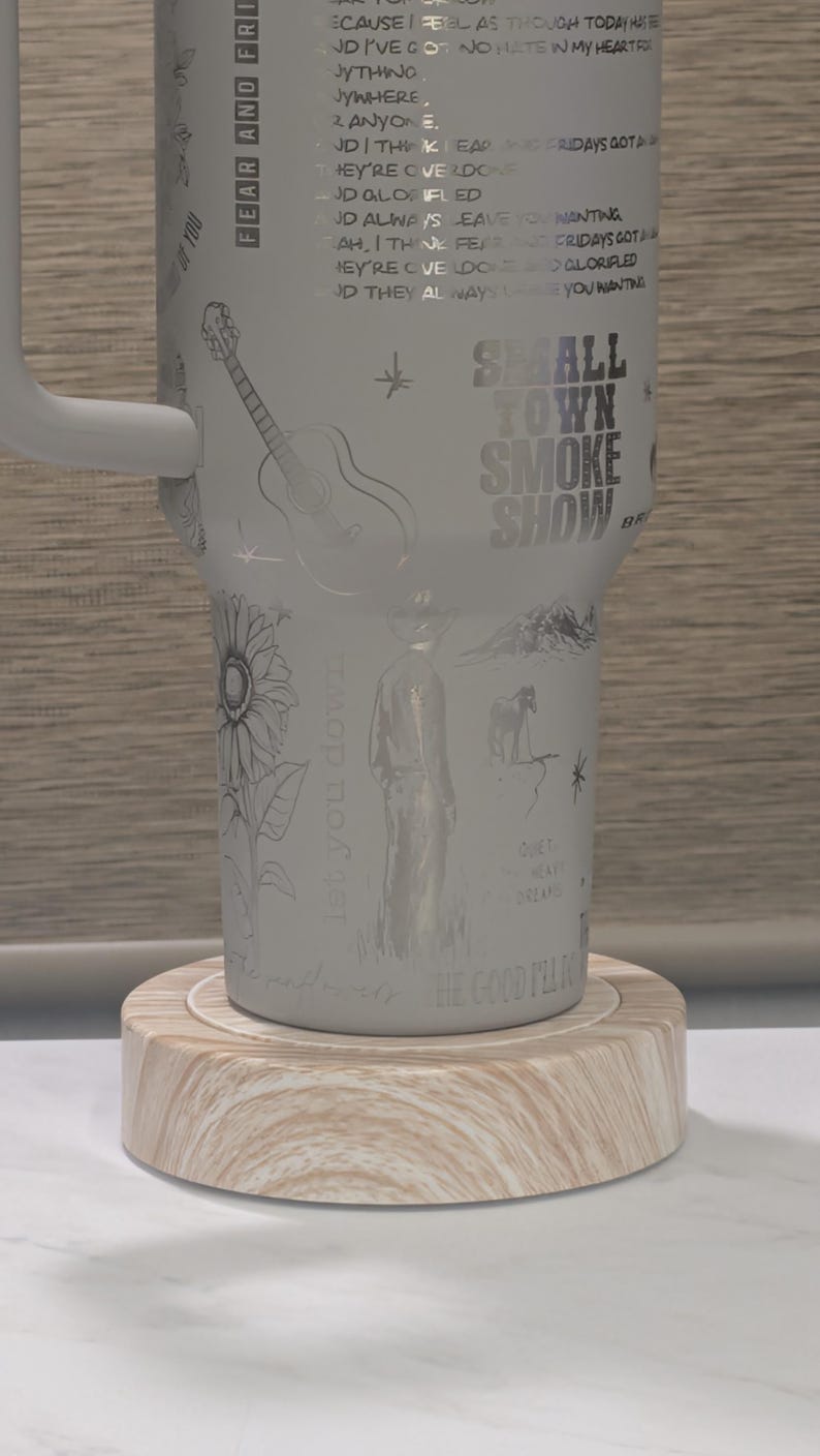 Może przedstawiać: Biały kubek z uchwytem, z wytrawionymi ilustracjami gitary, słonecznika, kowboja i konia. Widoczny jest napis "SMALL TOWN SMOKE SHOW". Kubek stoi na drewnianej podstawce.